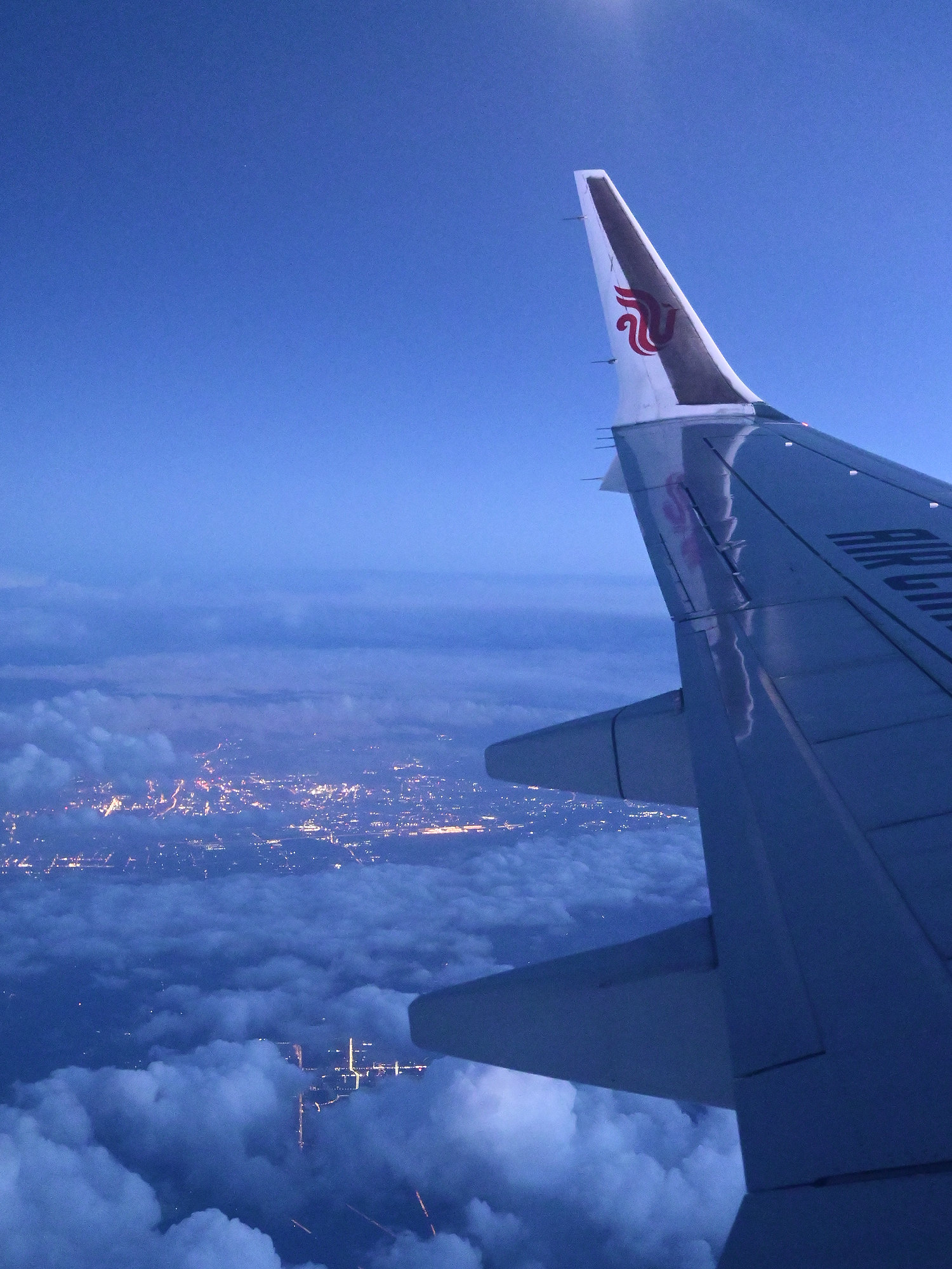 A moon above the aircraft wing on a night flight, with city lights visible beneath the clouds