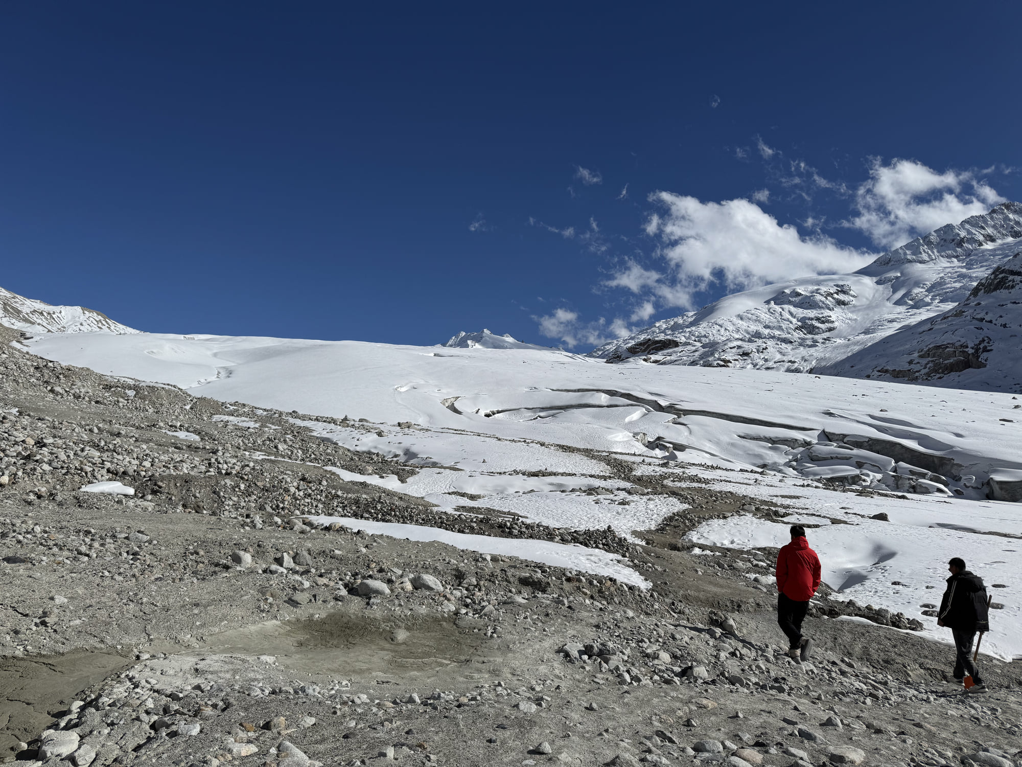 Two companions, seen from behind, walking across scree toward a snow-covered valley