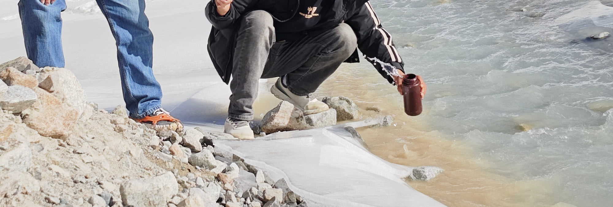 Hands, a container, and water during a two-person water-sampling operation beside snow and ice